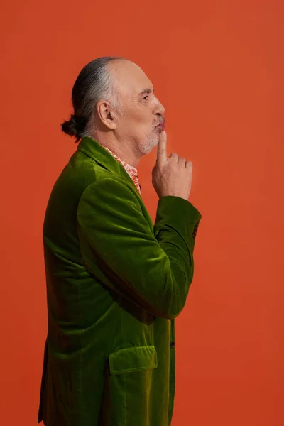 Vista lateral do homem sênior pensativo apontando com o dedo em lábios de puff ao pensar no fundo alaranjado vermelho, cabelo e barba cinzentos, estilo casual na moda, blazer de veludo verde, conceito de moda e idade — Fotografia de Stock