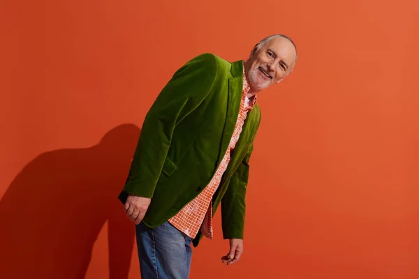 Charismatic and carefree senior model posing on red orange background with shadow and smiling at camera, trendy casual clothes, green velour blazer, personal style and positive aging concept — Stock Photo