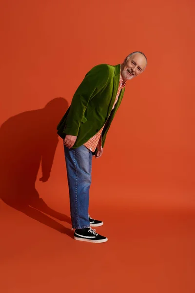 Homme âgé joyeux et barbu en vêtements décontractés tendance debout et regardant la caméra sur fond rouge orange, blazer en velours vert, jeans en denim bleu, concept de vieillissement heureux — Photo de stock