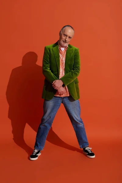 Volle Länge des coolen und selbstbewussten älteren Mannes in stylischer Freizeitkleidung stehend und in die Kamera blickend auf rot-orangefarbenem Hintergrund mit Schatten, grüner Velours-Blazer, blaue Jeans, Modelook — Stockfoto