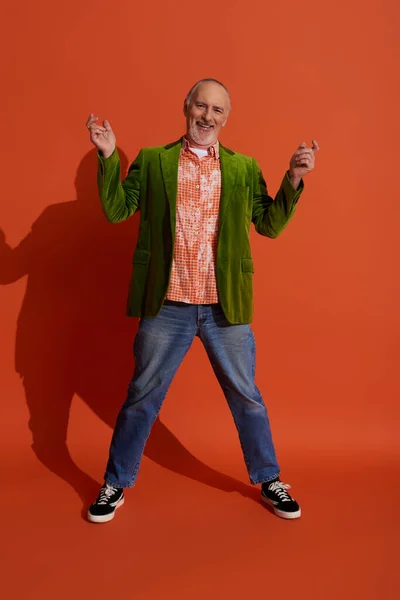 Glücklich alternder, lebensfroher, grauhaariger und bärtiger älterer Herr, der Spaß auf rot-orangefarbenem Hintergrund hat, grüner Velours-Blazer, trendiges Hemd, blaue Jeans, Mode- und Alterskonzept — Stockfoto