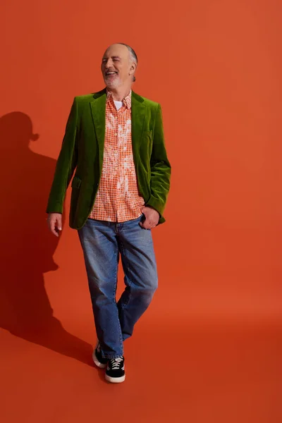 Homme senior positif en chemise tendance et blazer en velours vert posant avec pouce dans la poche de jeans en denim bleu tout en se tenant debout sur fond rouge orange, concept de vieillissement élégant — Photo de stock