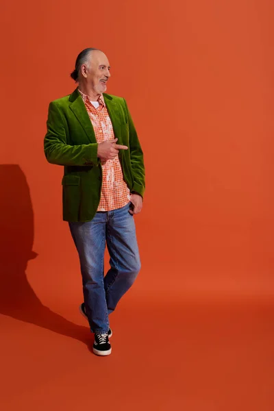 Piena lunghezza di uomo anziano fresco e allegro in giacca di velluto verde in piedi con mano in tasca di jeans di jeans blu, guardando altrove e puntando con il dito su sfondo arancione rosso con ombra — Foto stock