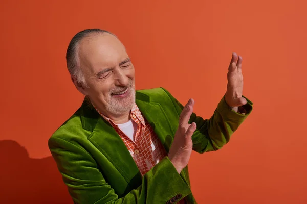 Excited grey haired and bearded senior man in green velour blazer showing stop gesture and laughing with closed eyes on red orange background, personal style, fashionable aging concept — Stock Photo
