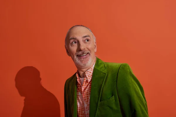 Joyful and surprised senior man with grey hair and beard smiling and looking away on red orange background, fashionable casual clothes, green velour blazer, trendy shirt, positive aging concept — Stock Photo