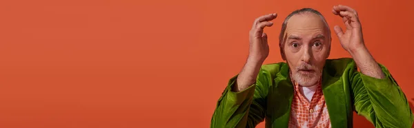 Homem idoso atordoado com cabelos grisalhos, barba e olhos salientes de mãos dadas perto da cabeça e olhando para a câmera no fundo laranja vermelho, blazer de veludo verde na moda, estilo pessoal, banner com espaço de cópia — Fotografia de Stock