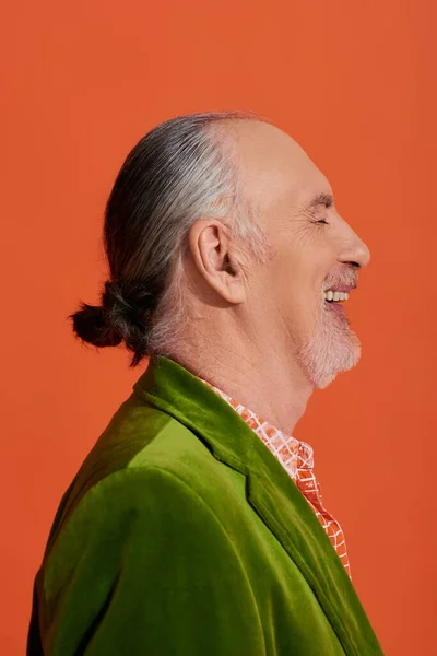 Side view of excited and fashionable older model laughing with closed eyes on vibrant orange background, portrait of cheerful and bearded senior man in green velour blazer — Stock Photo