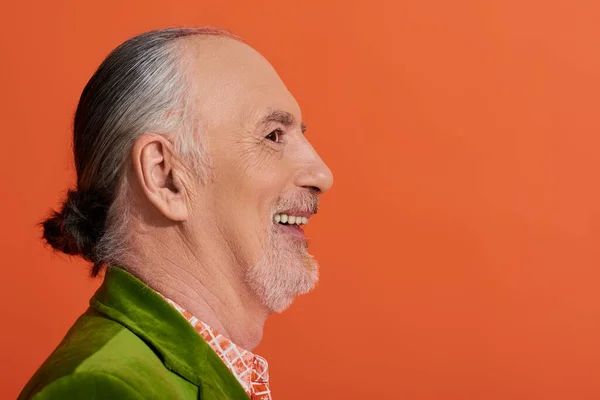 Профільний портрет харизматичної і радісної старої моделі, посміхаючись на яскравому помаранчевому фоні, старший чоловік з сірим волоссям і бородою, одягнений в зелений велюровий блейзер, щаслива і стильна концепція старіння — стокове фото