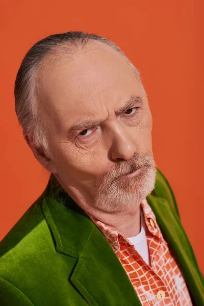 Porträt eines beleidigten älteren Mannes mit missmutigem Gesichtsausdruck, der auf leuchtend orangefarbenem Hintergrund in die Kamera blickt, älteres Modell, graue Haare, bärtiger, grüner Velours-Blazer, modisches Alterskonzept — Stockfoto