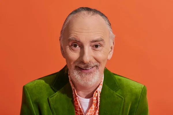 Retrato de modelo masculino senior carismático con pelo gris, barba y sonrisa radiante mirando a la cámara sobre fondo naranja vibrante, chaqueta de terciopelo verde, ropa casual de moda, envejecimiento positivo - foto de stock