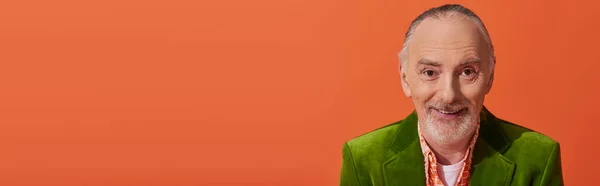 Retrato de modelo senior de moda y alegre con pelo gris y barba, vistiendo chaqueta de terciopelo verde y sonriendo a la cámara sobre fondo naranja vibrante, concepto de moda y edad, pancarta con espacio para copiar — Stock Photo