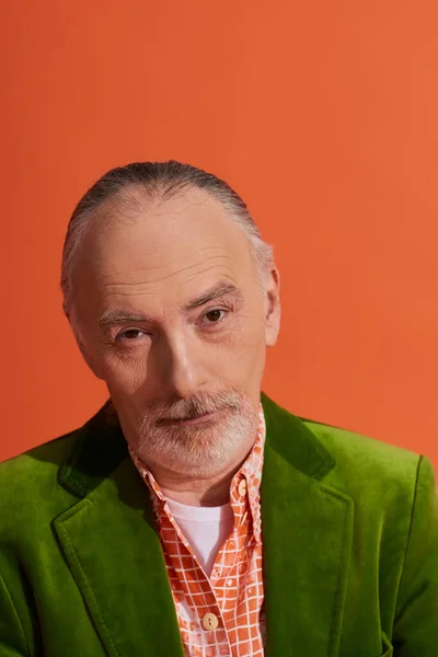 Retrato de escéptico y reflexivo hombre mayor de pelo gris, barbudo con chaqueta de terciopelo verde y mirando a la cámara en un fondo naranja vibrante, moda casual, concepto de envejecimiento de moda - foto de stock