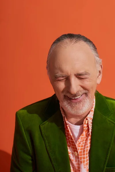 Happiness, portrait of overjoyed senior and bearded man in green velour blazer laughing with closed eyes on vibrant orange background, fashion look, positive and stylish aging concept — Stock Photo
