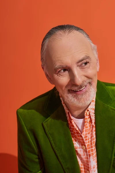 Stylish aging and positive lifestyle concept, portrait of optimistic senior male model smiling and looking away on vibrant orange background, grey hair, groomed beard, green velour blazer — Stock Photo