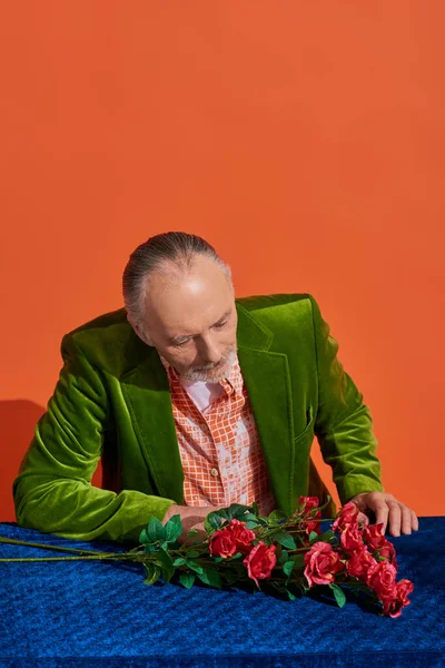 Melancolía, recuerdos, hombre mayor reflexivo en blazer de terciopelo verde y de moda mirando rosas rojas frescas en tela de terciopelo azul mientras está sentado sobre un fondo naranja vibrante con espacio para copiar - foto de stock