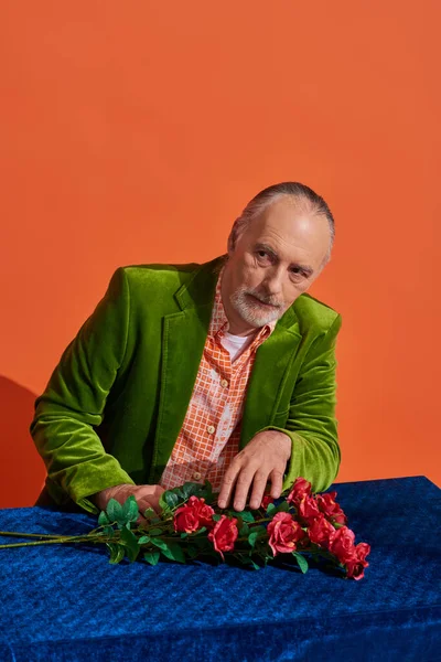 Anciano hombre reflexivo sentado cerca de la mesa con tela azul y rosas rojas mientras mira hacia otro lado sobre fondo naranja vibrante, modelo barbudo senior en chaqueta de terciopelo verde, concepto de envejecimiento de moda - foto de stock