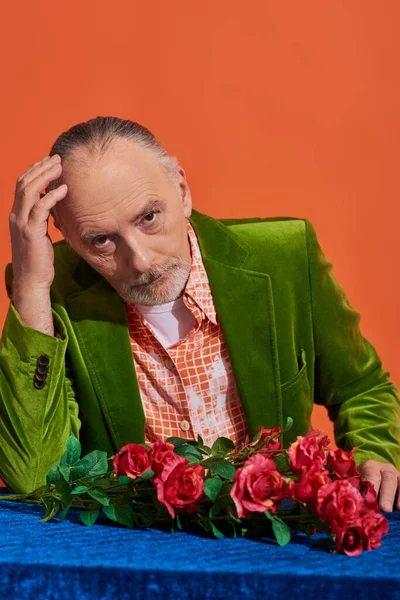 Pensive and serious senior man sitting near red roses on table with blue cloth, touching head and looking at camera on vibrant orange background, green velvet blazer, fashion and age concept — Stock Photo