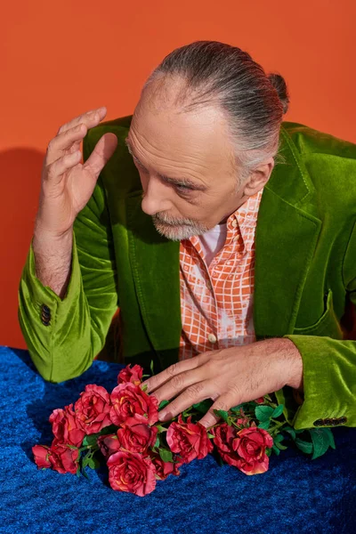 Gentiluomo anziano premuroso guardando altrove e seduto vicino a rose rosse su un panno di velluto blu su uno sfondo arancione vivace, capelli grigi e barba, giacca di velluto verde, stile personale, concetto di invecchiamento elegante — Foto stock