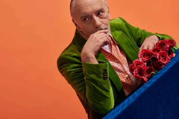Trendy older model in green velvet blazer, senior and pensive man holding hand near face while sitting next to red roses on blue velour cloth and looking away on vibrant orange background — Stock Photo