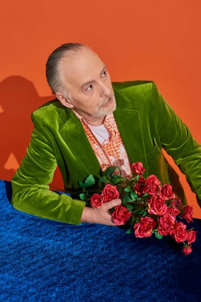 Thoughtful and fashionable senior man in green velvet blazer sitting with bouquet of red roses near table with blue velour cloth and looking away on vibrant orange background, stylish aging concept — Stock Photo