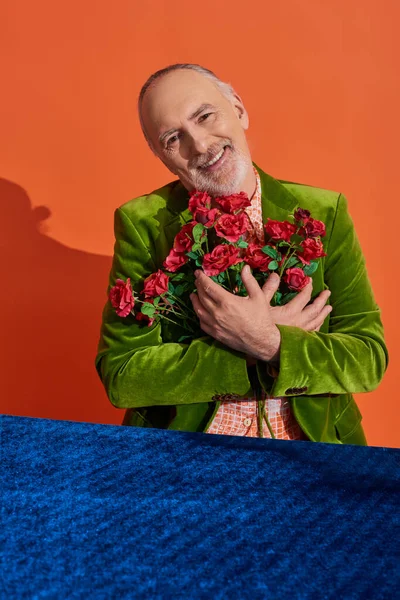 Modelo masculino senior lleno de alegría en chaqueta de terciopelo verde abrazando ramo de rosas rojas y sonriendo a la cámara cerca de la mesa con tela de terciopelo azul sobre fondo naranja vibrante, concepto de envejecimiento feliz - foto de stock