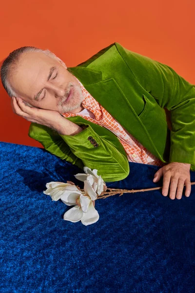 Glückliche Erinnerungen, älterer Herr sitzt neben weißer Orchidee auf blauem Velourtisch und lächelt mit geschlossenen Augen auf leuchtend orangefarbenem Hintergrund, grünem Samtblazer, positivem Alterungskonzept, Blickwinkel hoch — Stockfoto