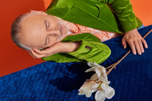 Glückliche Erinnerungen, Draufsicht auf lächelnden älteren Mann im grünen Samtblazer, der mit geschlossenen Augen neben weißer Orchidee auf Tisch mit blauem Velourtuch auf leuchtend orangefarbenem Hintergrund sitzt, positives Alterungskonzept — Stockfoto