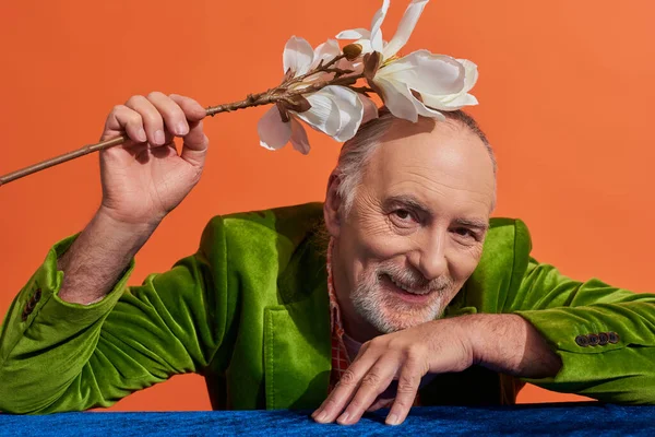 Happy aging concept, delighted and bearded senior man in green velvet blazer sitting at table with blue velour cloth and holding white orchid flower above head on vibrant orange background — Stock Photo