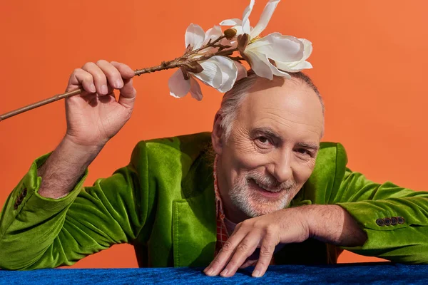 Conceito de envelhecimento feliz, homem sênior encantado e barbudo em blazer de veludo verde sentado à mesa com pano de veludo azul e segurando flor de orquídea branca acima da cabeça no fundo laranja vibrante — Fotografia de Stock