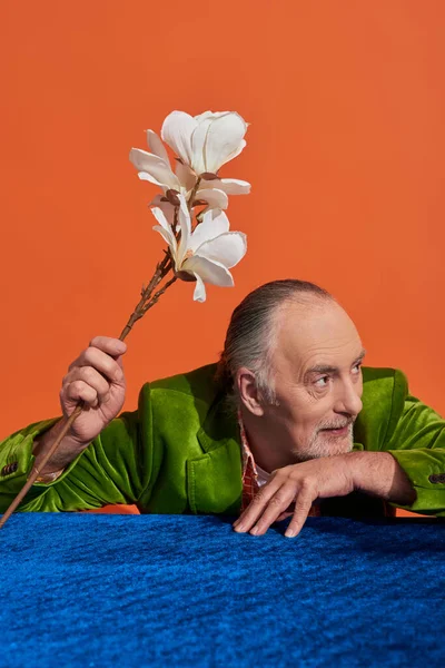 Joyful and grey haired senior man sitting at table with blue velour cloth, holding white orchid flower and looking away on vibrant orange background, green velvet blazer, fashion and age concept — Stock Photo