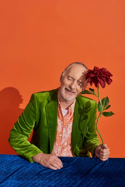 Pleased elderly and bearded man smiling with closed eyes while sitting near red peony and table with blue velour cloth on vibrant orange background, green velvet jacket, groomed beard, stylish aging — Stock Photo