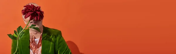 Emoción feliz, hombre mayor sonriente oscureciendo la cara con flor de peonía roja y sonriendo sobre un fondo naranja vibrante, chaqueta de terciopelo verde, concepto de envejecimiento elegante y positivo, pancarta con espacio para copiar — Stock Photo