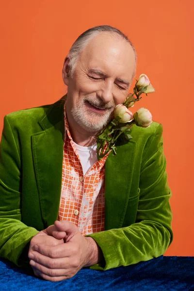 Zufriedener älterer Mann mit grauen Haaren, Bart und geschlossenen Augen, der mit frischen Rosen posiert und am Tisch mit blauem Velourtuch auf leuchtend orangefarbenem Hintergrund lächelt, grüner Samtblazer, stylisches Alterskonzept — Stockfoto
