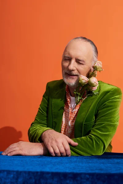 Pleased aged man smiling with closed eyes while sitting with fresh roses near table with blue velour cloth on vibrant orange background, green velvet blazer, person style, happy aging concept — Stock Photo