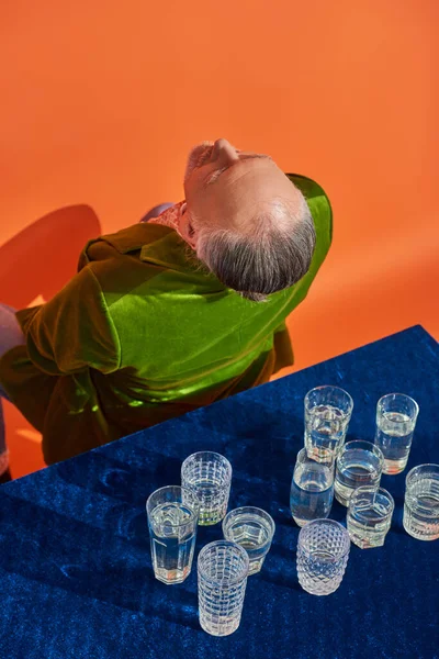 Vista dall'alto dell'uomo anziano in giacca di velluto verde seduto con schiena a tavola con panno di velluto blu e bicchieri d'acqua su sfondo arancione, invecchiamento della popolazione, simbolismo, concetto di pienezza di vita — Foto stock