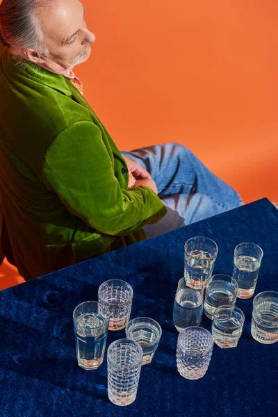 Стильний і продуманий старший чоловік у зеленому оксамитовому блістері, що сидить біля окулярів зі свіжою чистою водою на столі з синьою велюровою тканиною, символікою, концепцією повноти життя, високим кутом зору — стокове фото