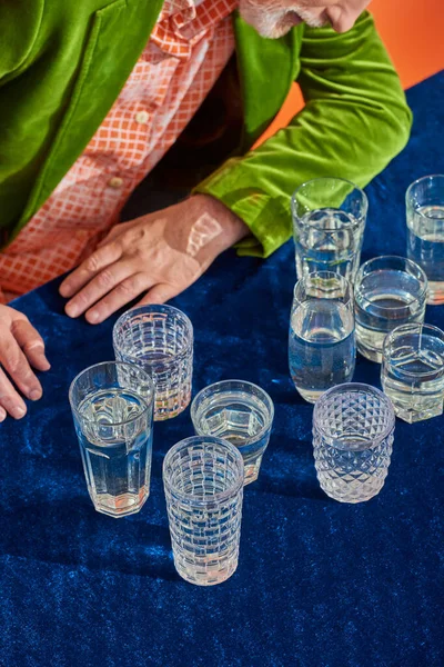Cropped view of senior man in trendy and green velvet blazer sitting near crystal glasses with clear water on table with blue velour cloth on orange background, symbolism, life fullness concept — Stock Photo