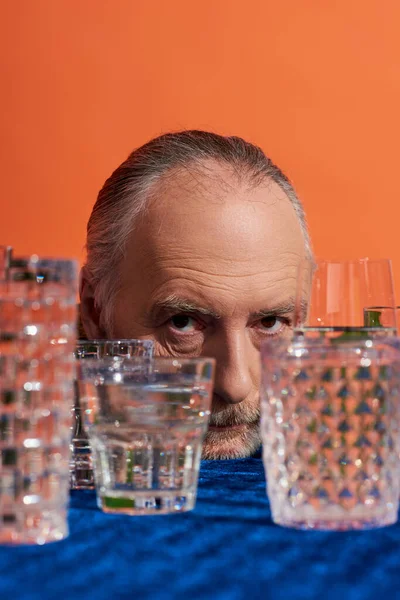 Portrait d'un homme aux cheveux gris âgé au regard expressif regardant une caméra près de verres en cristal remplis d'eau claire sur fond orange, population vieillissante, symbolisme, concept de plénitude de vie — Photo de stock