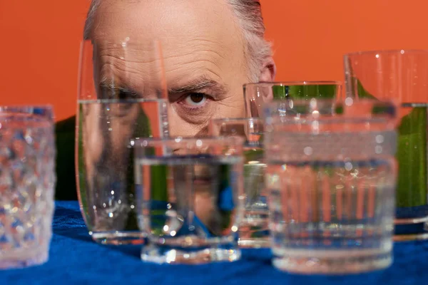 Modelo masculino senior con mirada expresiva mirando a la cámara detrás de vidrios de cristal borrosos con agua pura sobre fondo naranja, población envejecida, simbolismo, concepto de plenitud de vida - foto de stock