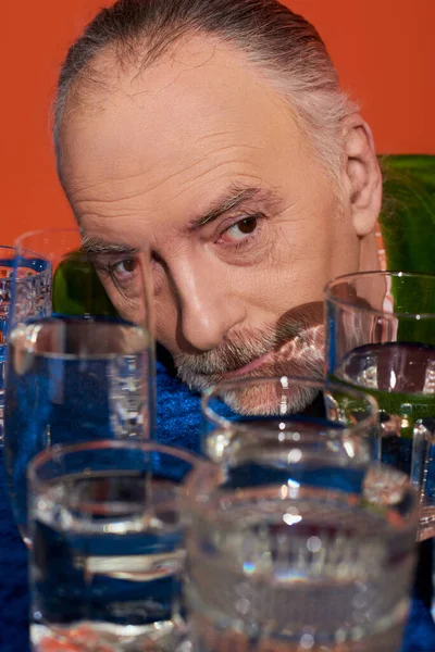 Erinnerungen, älterer und nachdenklicher Mann, der in der Nähe von Gläsern mit klarem Wasser auf einem Tisch mit blauem Velourtuch auf orangefarbenem Hintergrund wegschaut, alternde Bevölkerung, Symbolik, Lebensfüllungskonzept — Stockfoto
