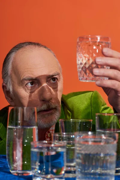 Älterer und nachdenklicher grauhaariger Mann in grünem Samtblazer mit Blick auf ein Glas voll reinen Wassers am Tisch mit blauem Tuch auf orangefarbenem Hintergrund, alternde Bevölkerung, Symbolik, Lebensfüllungskonzept — Stockfoto