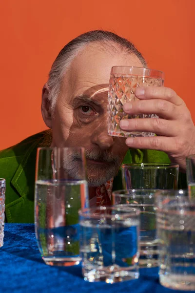 Älterer grauhaariger Mann in grünem Samtblazer, der sein Gesicht mit einem Glas reinen Wassers verdeckt und auf orangefarbenen Hintergrund in die Kamera blickt, alternde Bevölkerung, Symbolik, Konzept der Lebensfülle — Stockfoto