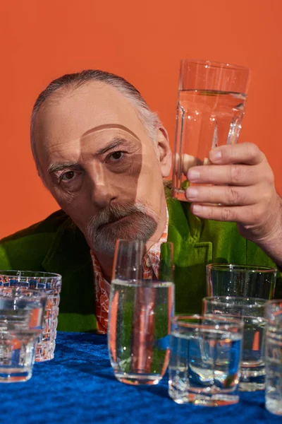 Senior bearded man in fashionable and green velvet blazer holding glass of clear water and looking at camera on orange background, aging population, symbolism, life fullness concept — Stock Photo