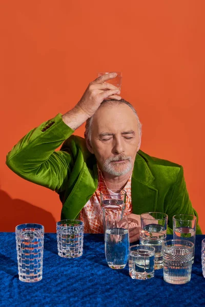 Homme âgé barbu avec les yeux fermés, dans un blazer de velours élégant et vert, tenant un verre d'eau au-dessus de la tête sur fond orange, vieillissement de la population, symbolisme, concept de plénitude de vie — Photo de stock