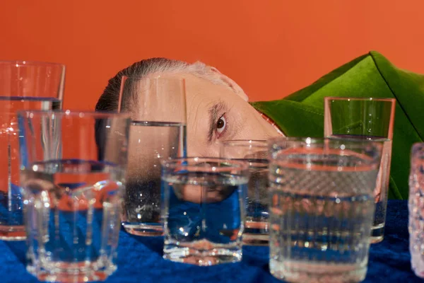 Пожилой человек скрывает лицо за прозрачными стаканами с чистой водой на столе с голубой велюровой тканью на оранжевом фоне, старение населения, символизм, концепция полноты жизни — стоковое фото