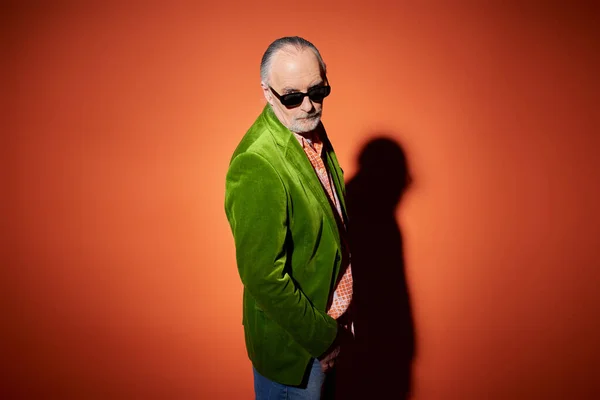 Senior male model in stylish casual clothes looking at camera on red and orange background with shadow, green velour blazer, dark sunglasses, expressive individuality, fashionable aging concept — Stock Photo