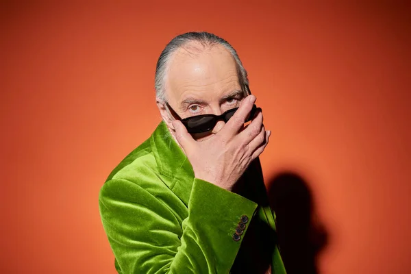 Anciano y hombre de moda en chaqueta de terciopelo verde quitándose las gafas de sol con estilo oscuro y mirando a la cámara con mirada expresiva sobre el fondo rojo y naranja, concepto de envejecimiento de moda — Stock Photo