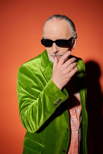 Hombre de edad avanzada de moda en gafas de sol oscuras y chaqueta de terciopelo verde posando con la mano cerca de la barba y mirando hacia otro lado sobre fondo rojo y naranja con sombra, moda y concepto de edad - foto de stock