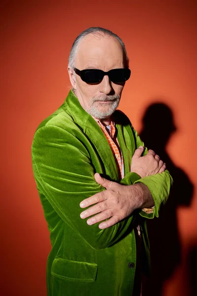 Homme âgé aux cheveux gris confiant et barbu debout avec les bras croisés sur fond rouge et orange avec ombre, lunettes de soleil sombres, veston en velours vert, personnalité vibrante, concept de vieillissement élégant — Photo de stock