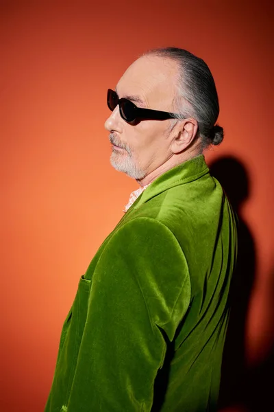 Вид збоку на сіроволосий і бородатий старший чоловік в темних сонцезахисних окулярах і зелений велюровий блейзер, що стоїть і дивиться на червоний і помаранчевий фон з тіньою, позитивною і модною концепцією старіння — стокове фото